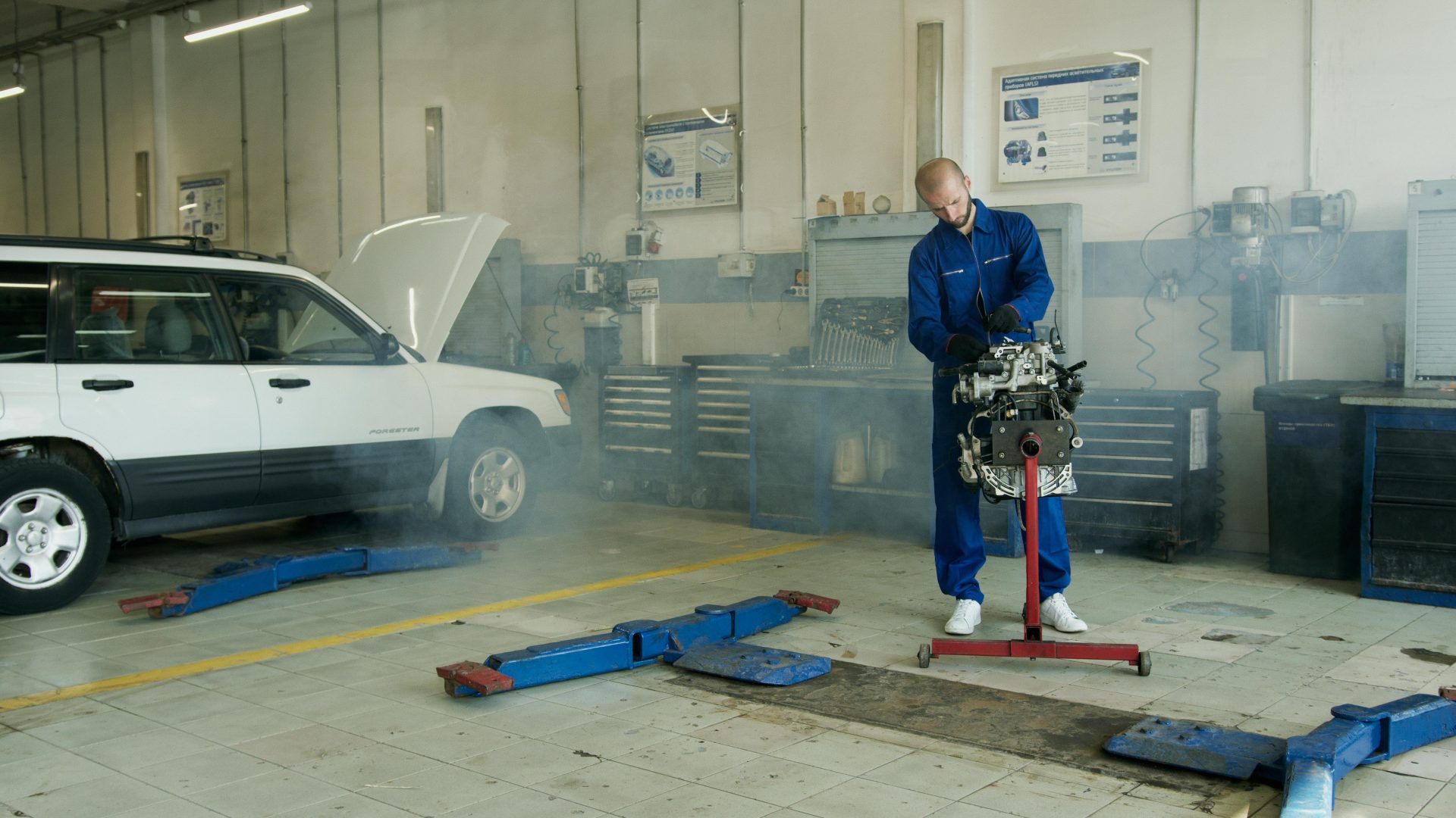 Mechaniker im blauen Overall arbeitet in einer Autowerkstatt an einem Automotor mit einem weißen Auto.